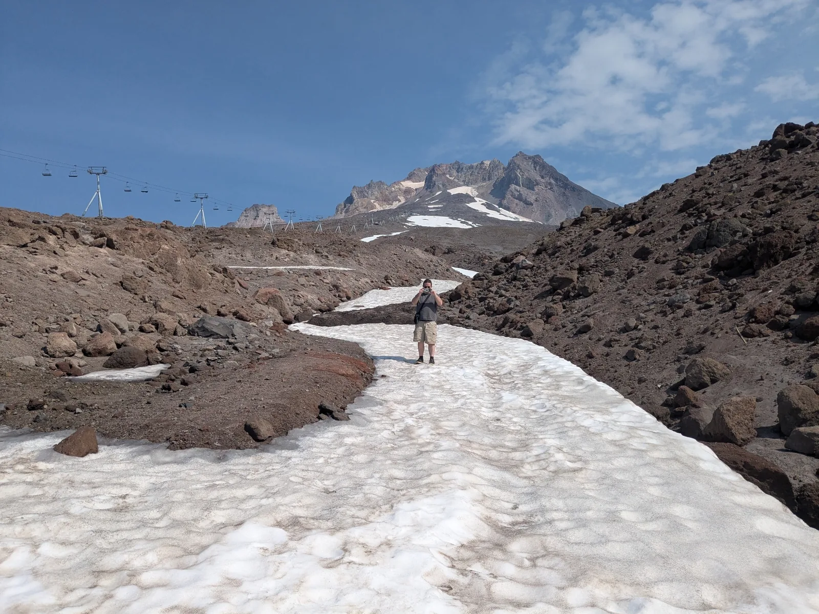 A hiker standing on a patch of snow below Mount Hood, surrounded by rocky slopes and ski lifts.