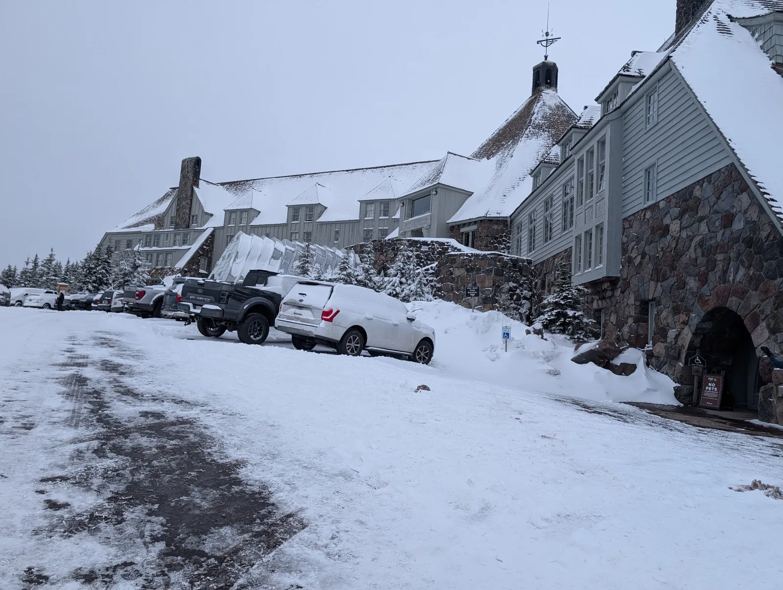 A mountain lodge with snow-covered roofs and cars parked along its stone exterior.